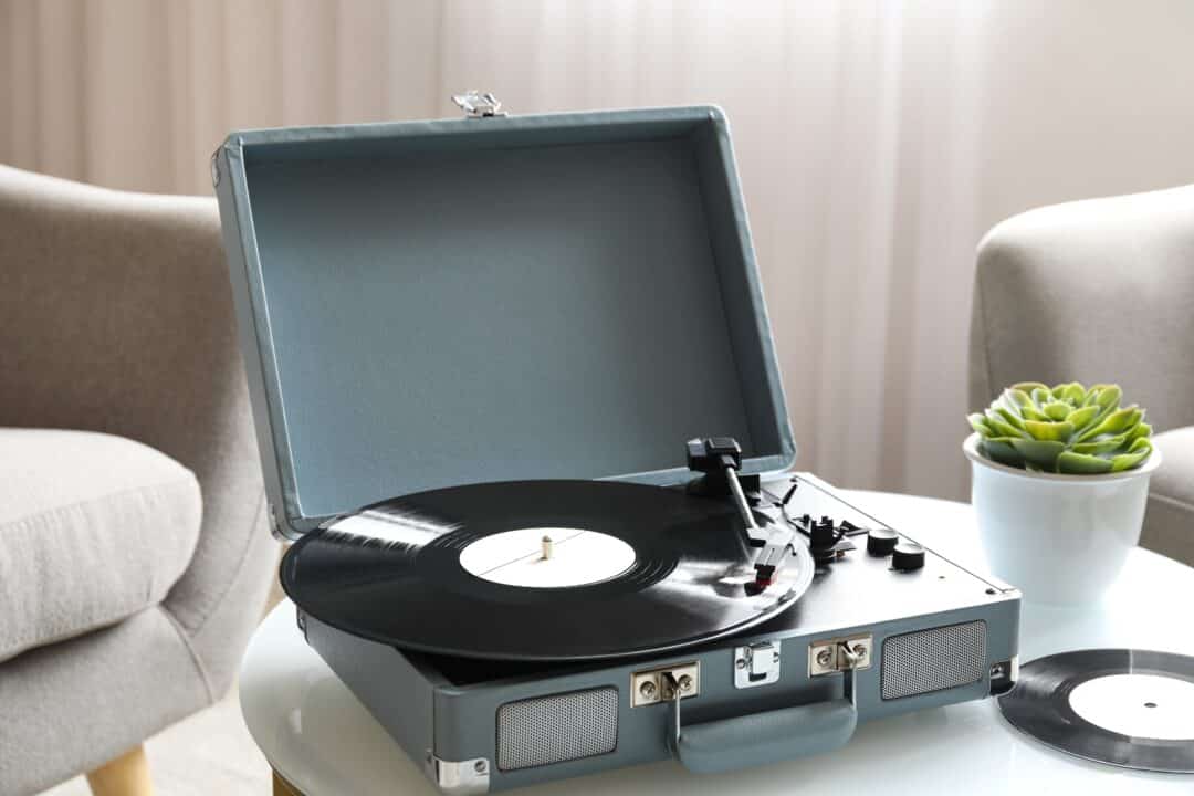 Bärbar vinylspelare på bord med krukväxt och fåtölj i bakgrunden.