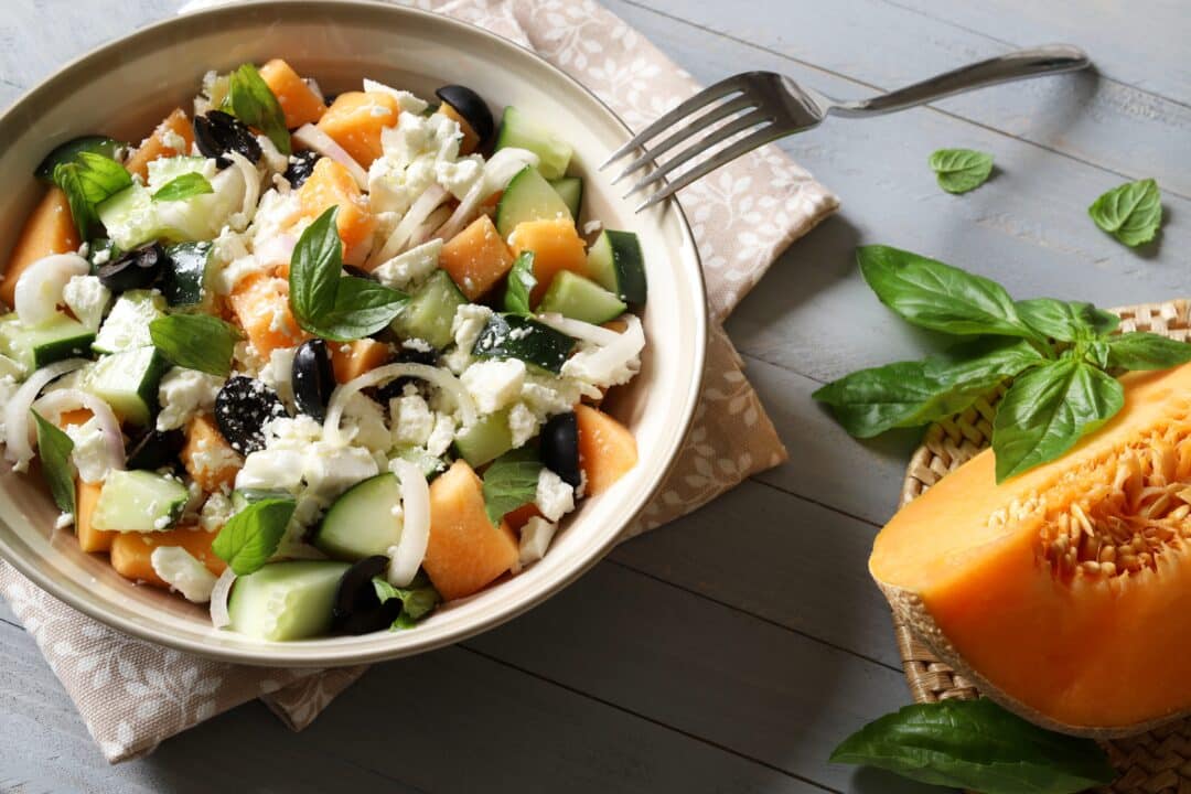 Frisk sommarsallad med melon, gurka, fetaost och oliver på ljus träbakgrund.