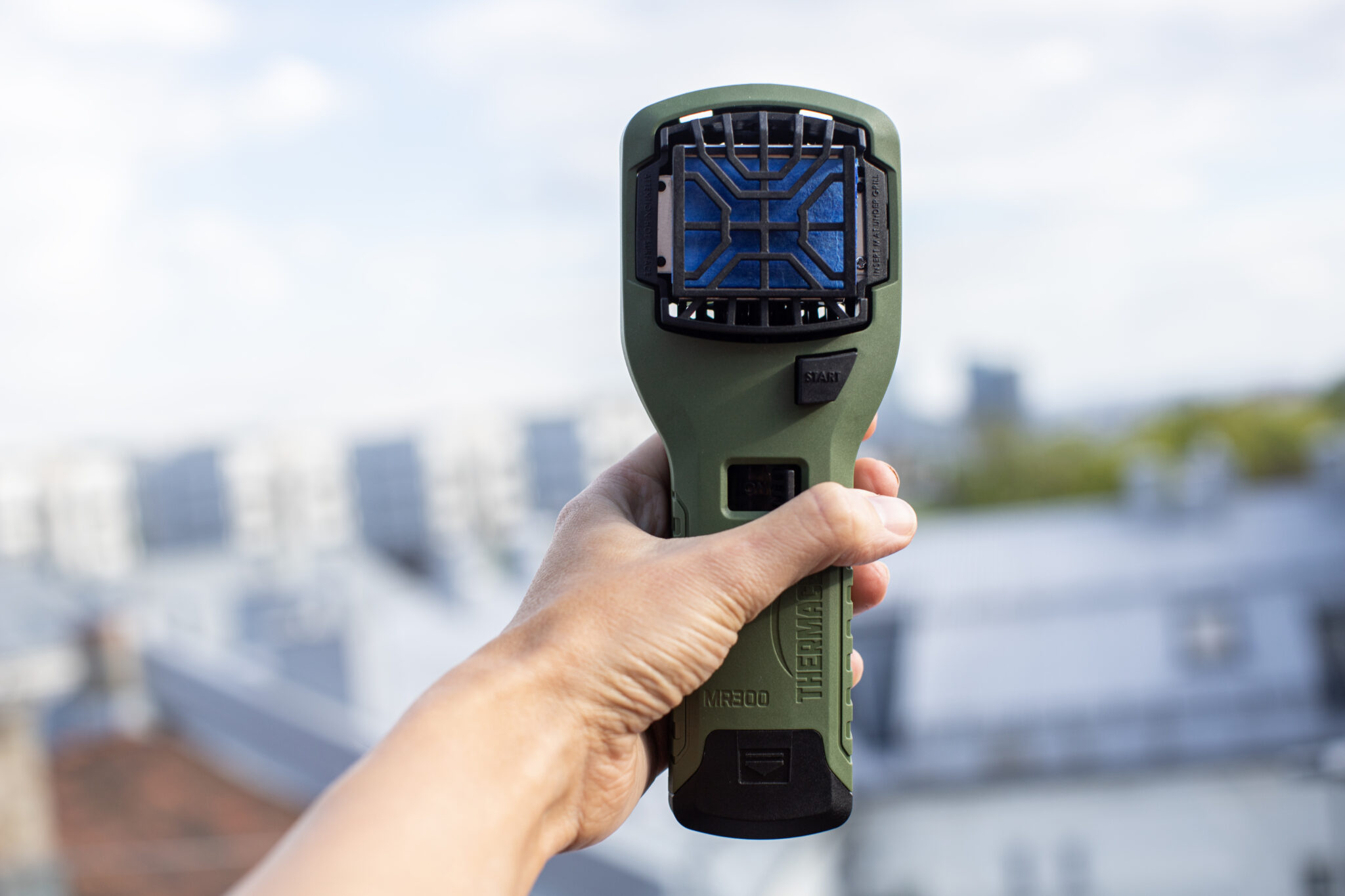 Handhållen thermometer med grön design, fotograferad med stadslandskap i bakgrunden.