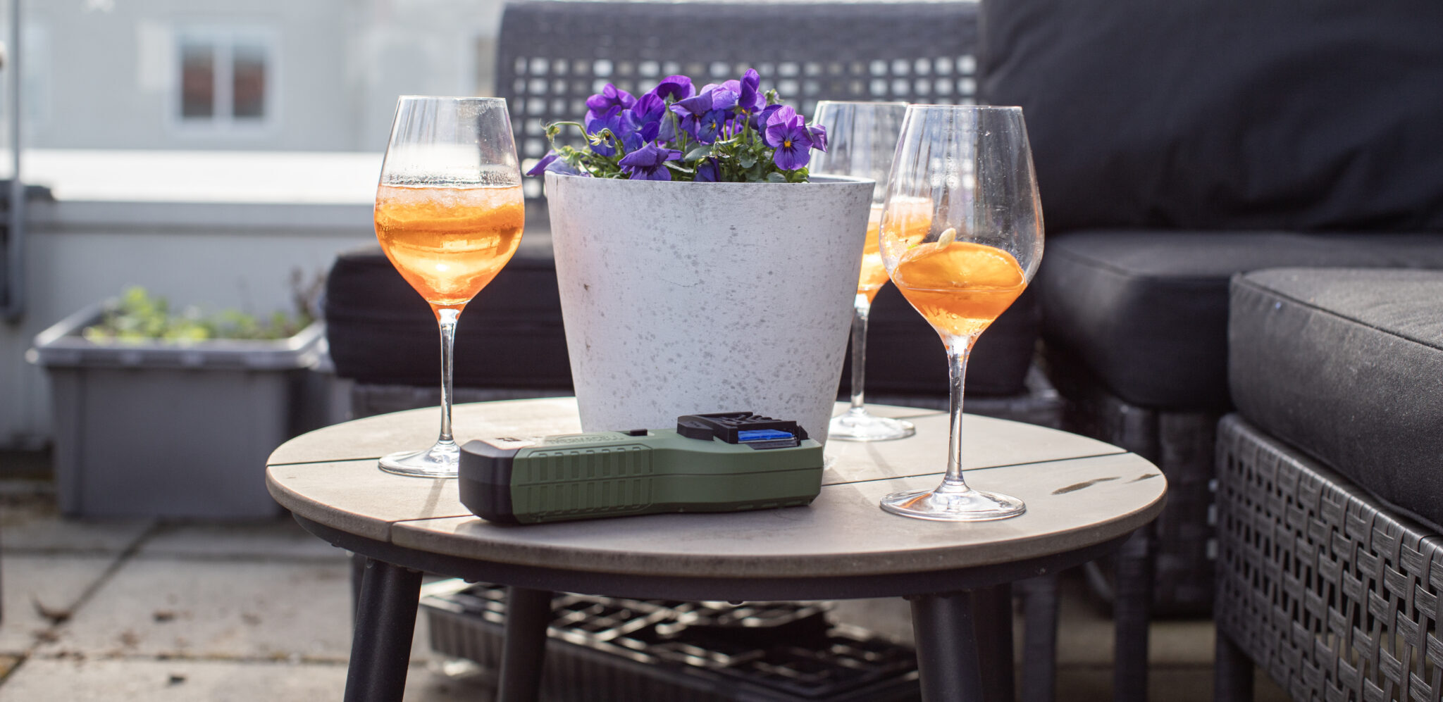 Alt-text: Tre drinkar på ett bord med blommor, en solig terrass och utemöbler.
